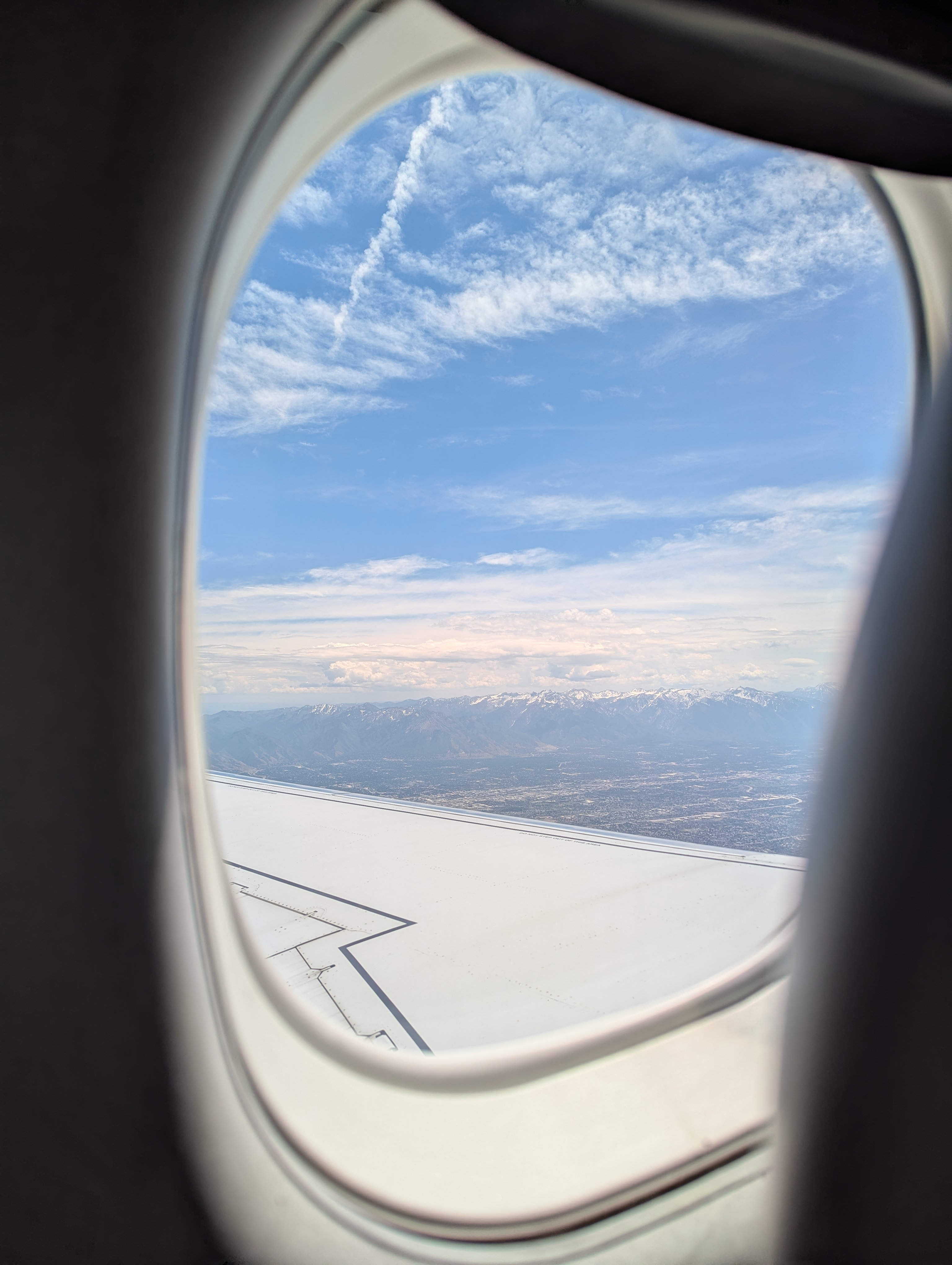 The camera is peeking through an airplane window. A snow-capped mountain range is in the background with the airplane wing in the foreground.