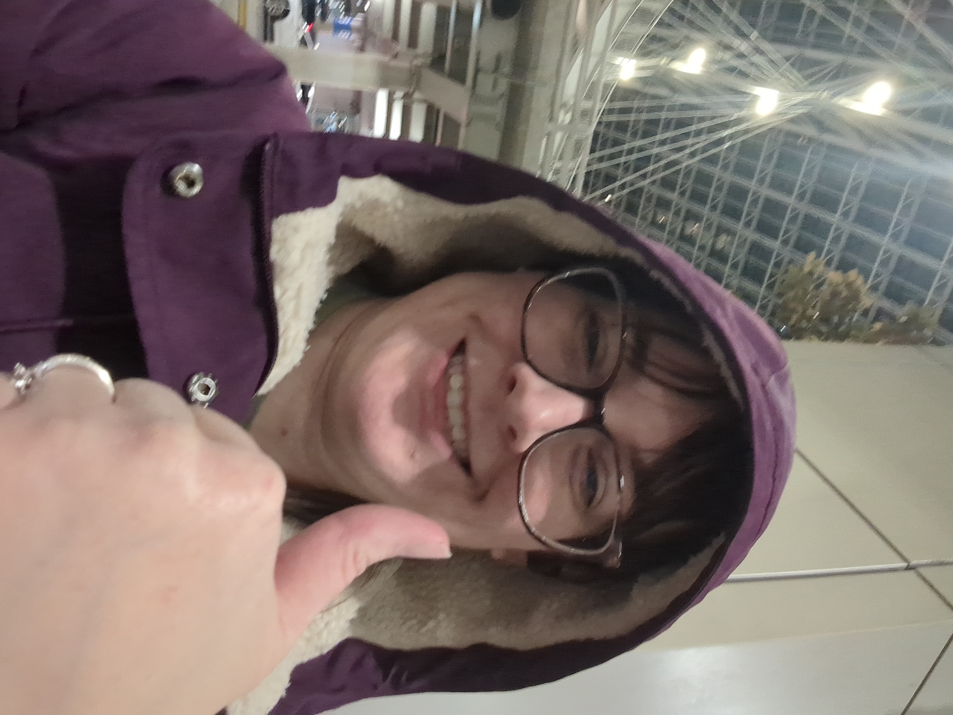 Kristen is sitting in front of an airport, looking tired and sick but smiling and giving a thumbs up